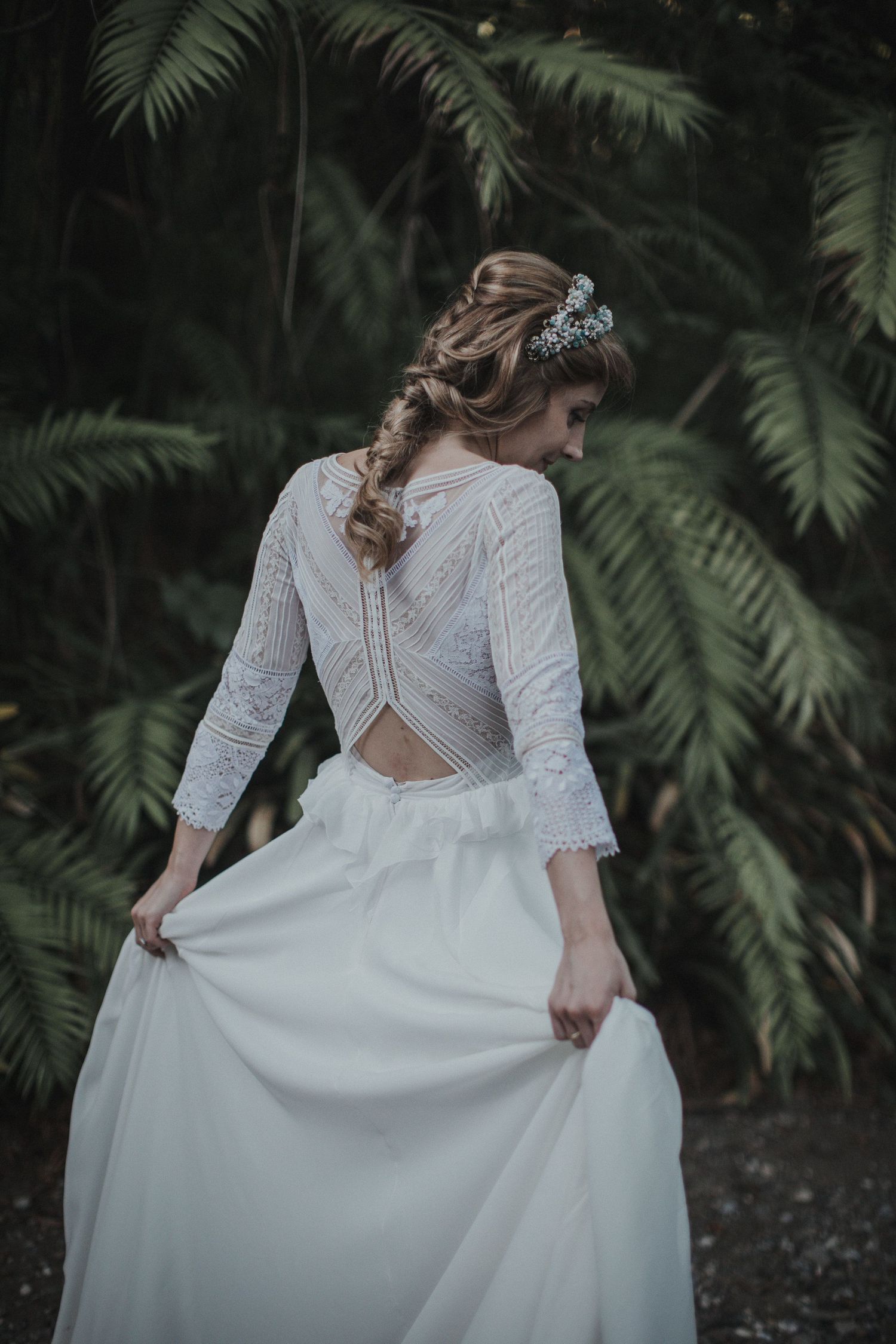 Bride in elegant white lace dress with braided hair poses showing intricate back detail against tropical foliage.
