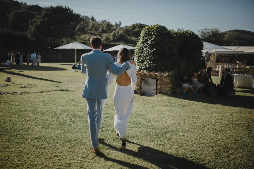 Couple in wedding attire walking away together across sunny lawn at outdoor reception.