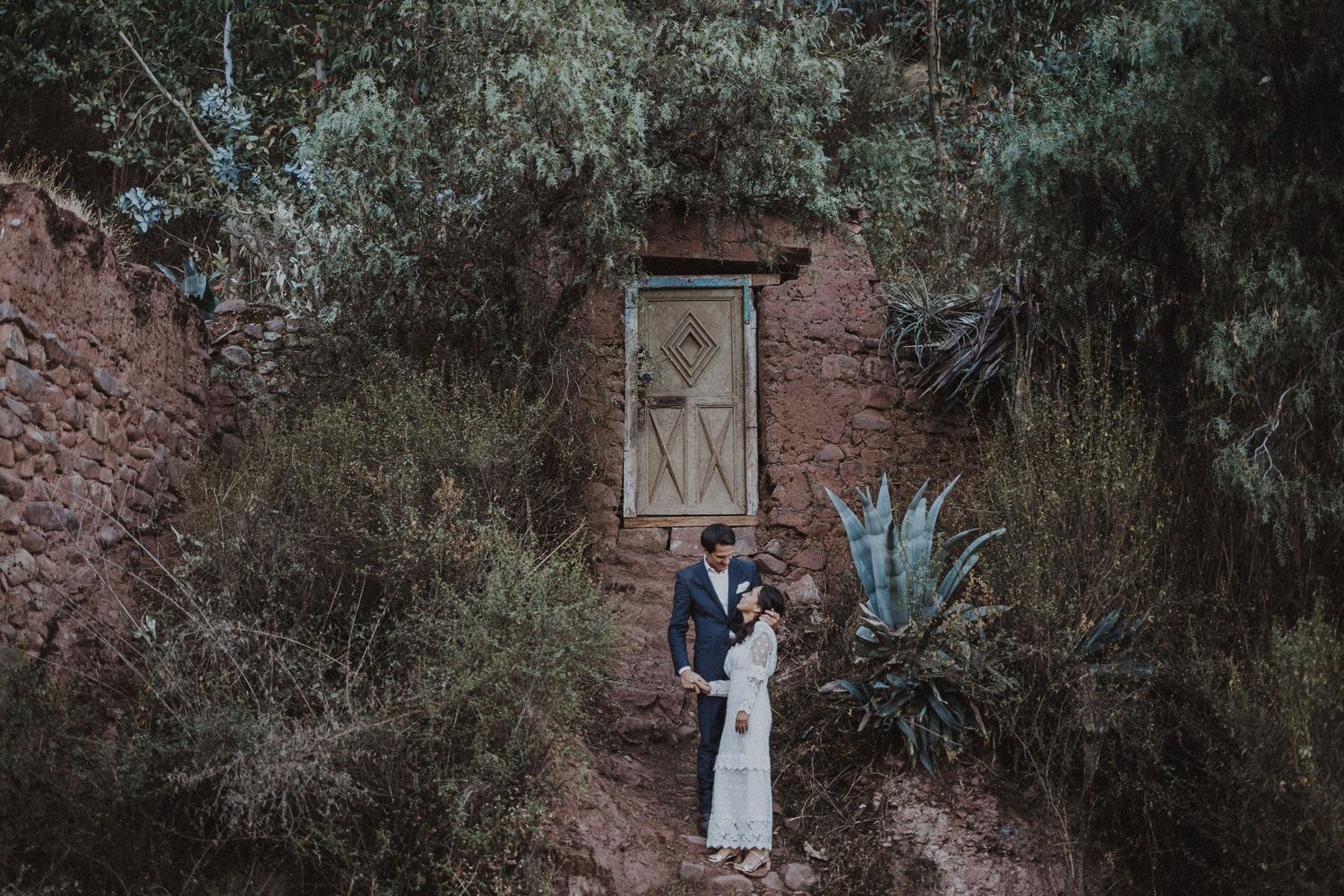 A couple stands together on stone steps near an ancient wall relief and blue agave plants.