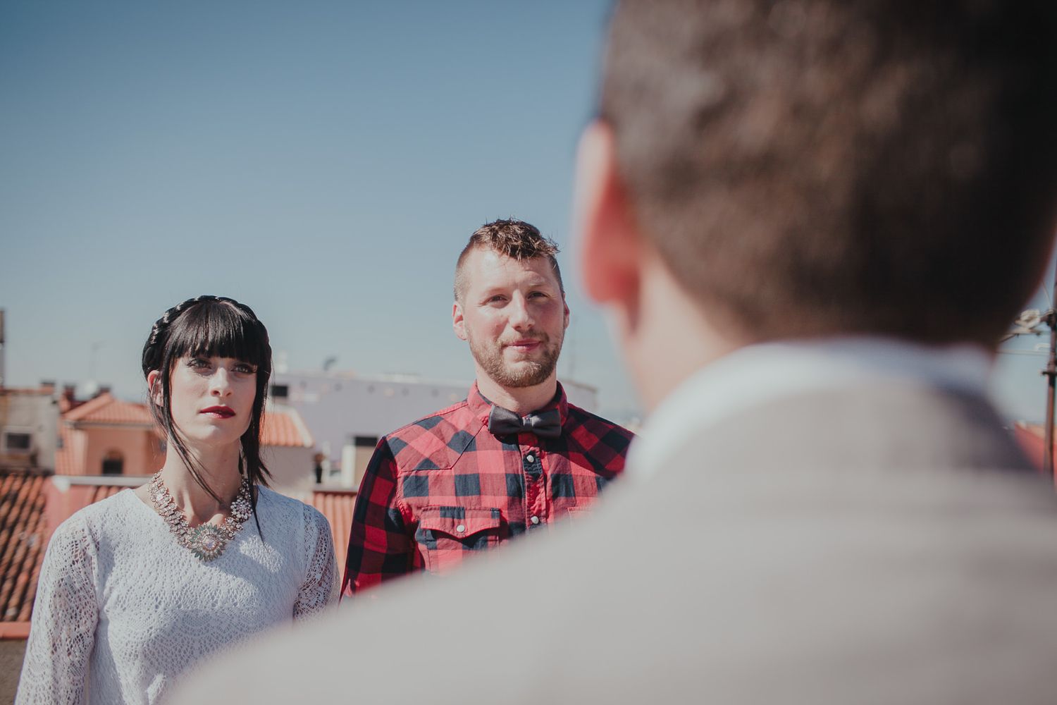 A couple and officiant stand on a rooftop terrace with Spanish clay tile buildings in the background on a sunny day.