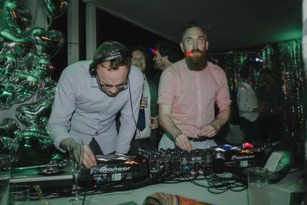 Two DJs working together at mixing decks under green party lighting.