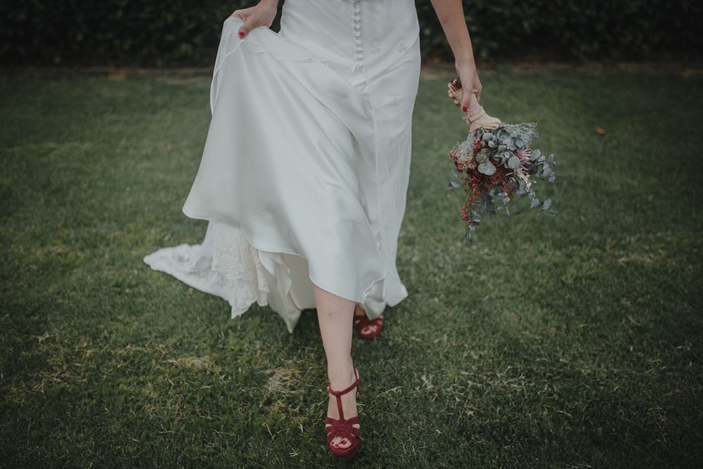 A close-up of red shoes and white dress hem while walking on grass with bouquet.