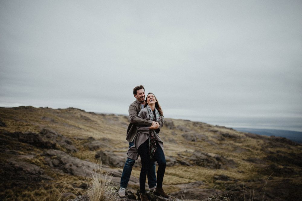 A couple embraces while standing on a rugged hillside overlooking a misty landscape on an overcast day.