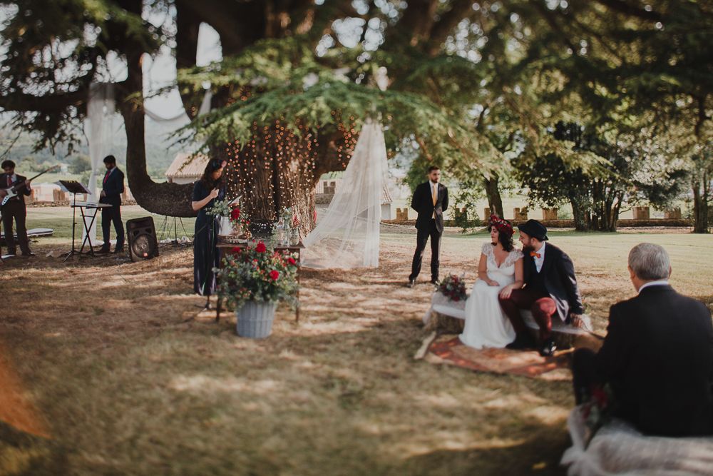 An outdoor wedding ceremony takes place under a decorated tree with hanging lights.