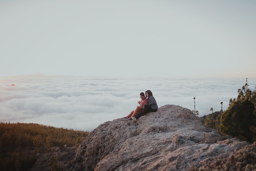 Two figures sit on rocky overlook above sea of clouds at sunset.