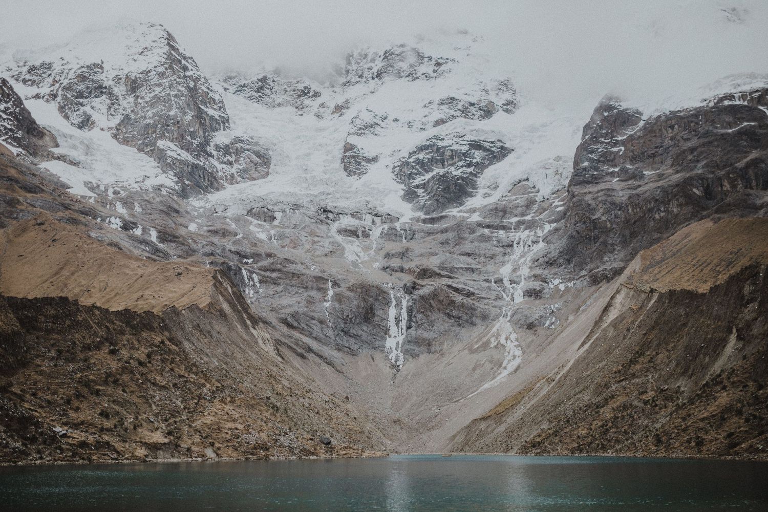 A glacial lake reflects snow-capped mountains and dramatic cliffs in a pristine wilderness setting.