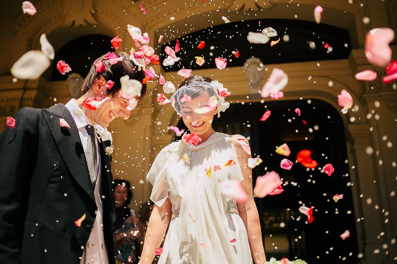 Jubilant wedding couple showered with rose petals against dramatic dark background.