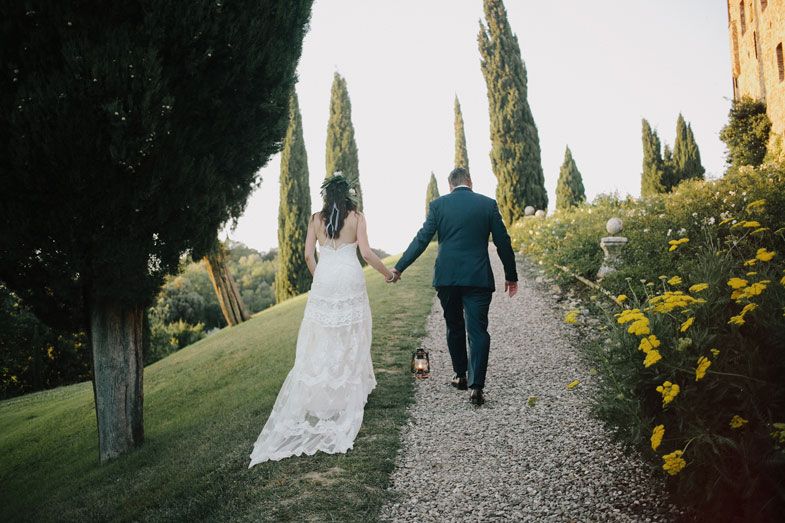 Newlyweds walking down a cypress-lined gravel path in an Italian garden setting.