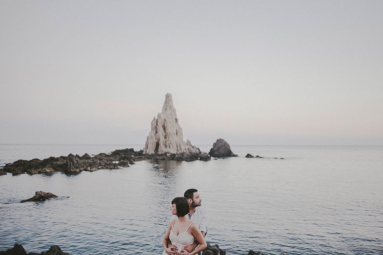Person sitting on rocky shore overlooking dramatic seaside rocks at dusk.