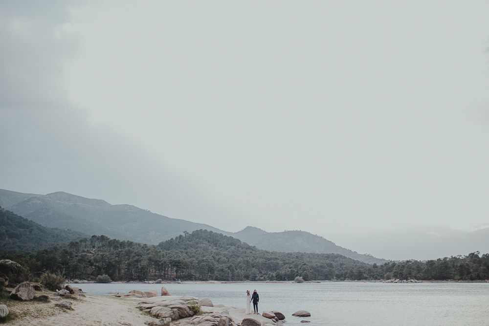 A lone figure stands on a rocky shoreline overlooking a misty mountain lake on an overcast day.