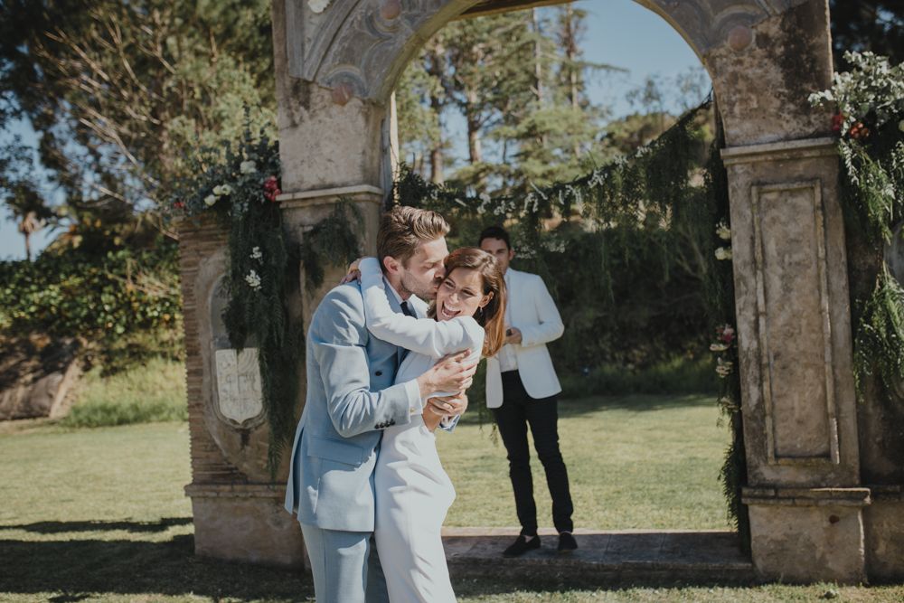 Newlyweds share first kiss under ornate stone archway during outdoor ceremony.