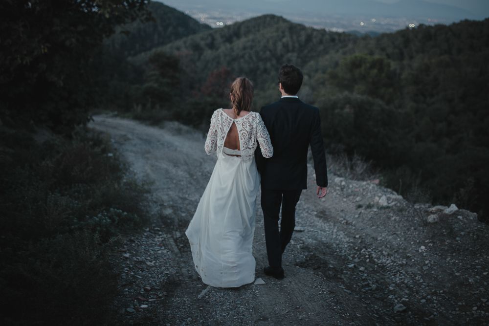 A couple walks away on a mountain path, showing off the bride's detailed lace back wedding dress.