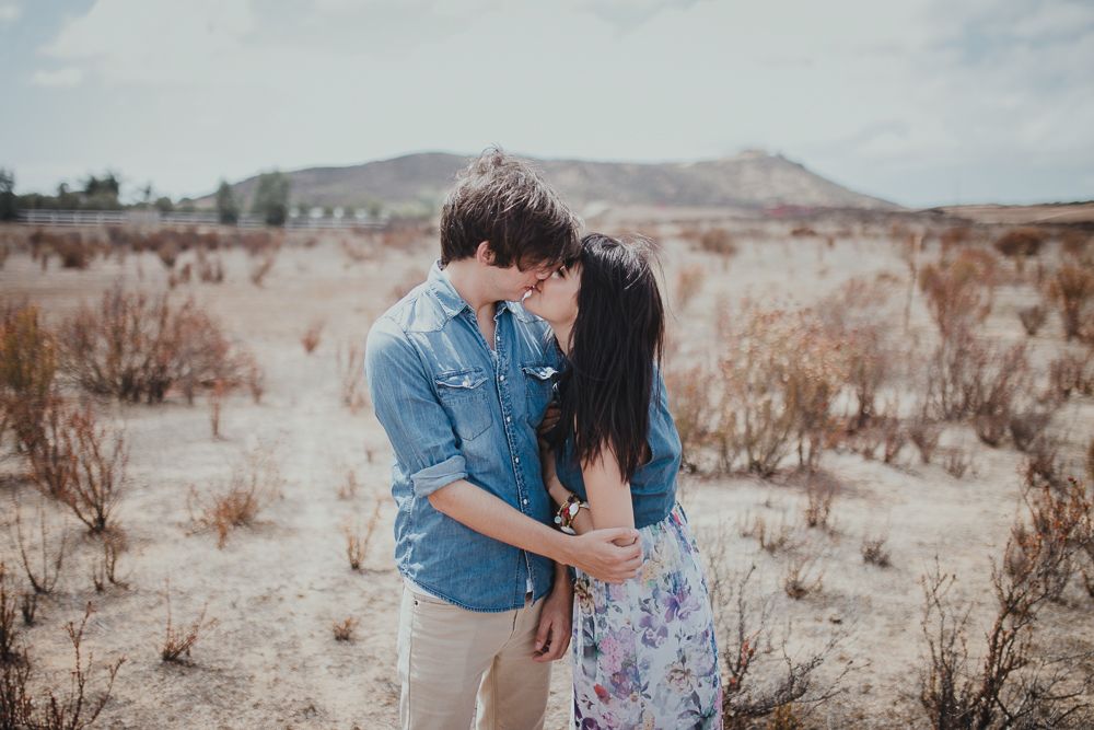 A couple shares an intimate moment among desert shrubs with mountains in the background.