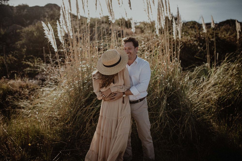 A romantic couple embraces in a field of tall dried grasses during sunset.