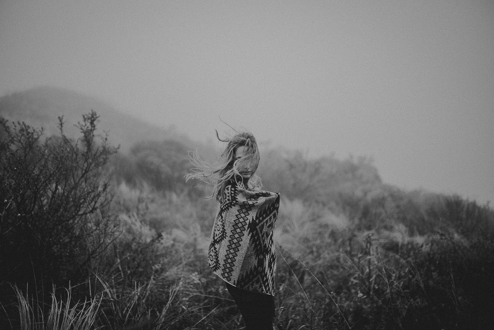 Figure stands alone in mountain landscape holding patterned blanket in black and white.