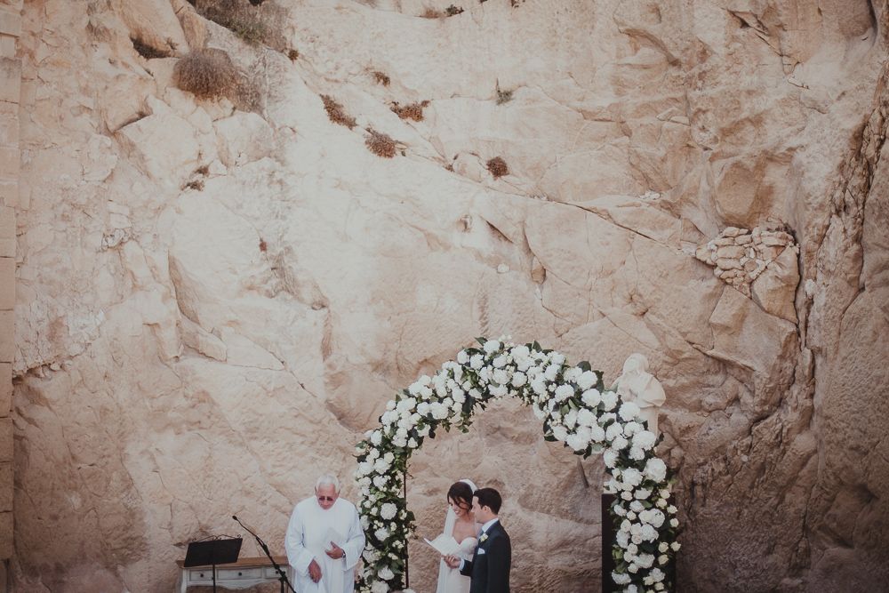 Wedding ceremony under a white floral arch against a rustic canyon wall.