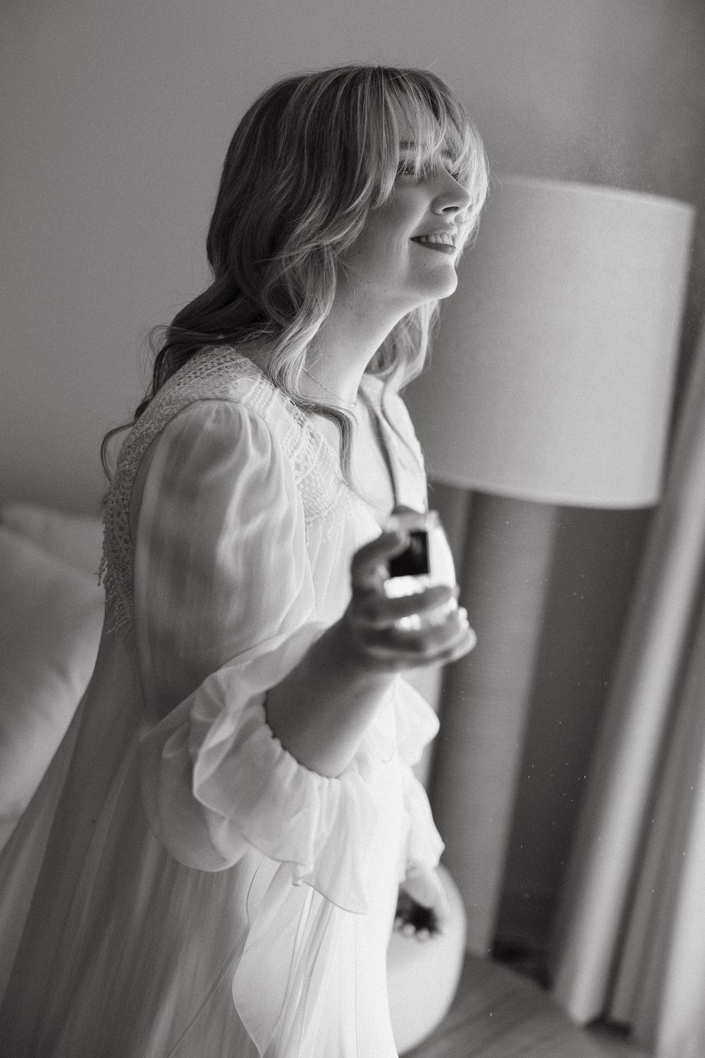 A woman in a flowing white dress smiles while getting ready in a sunlit room.