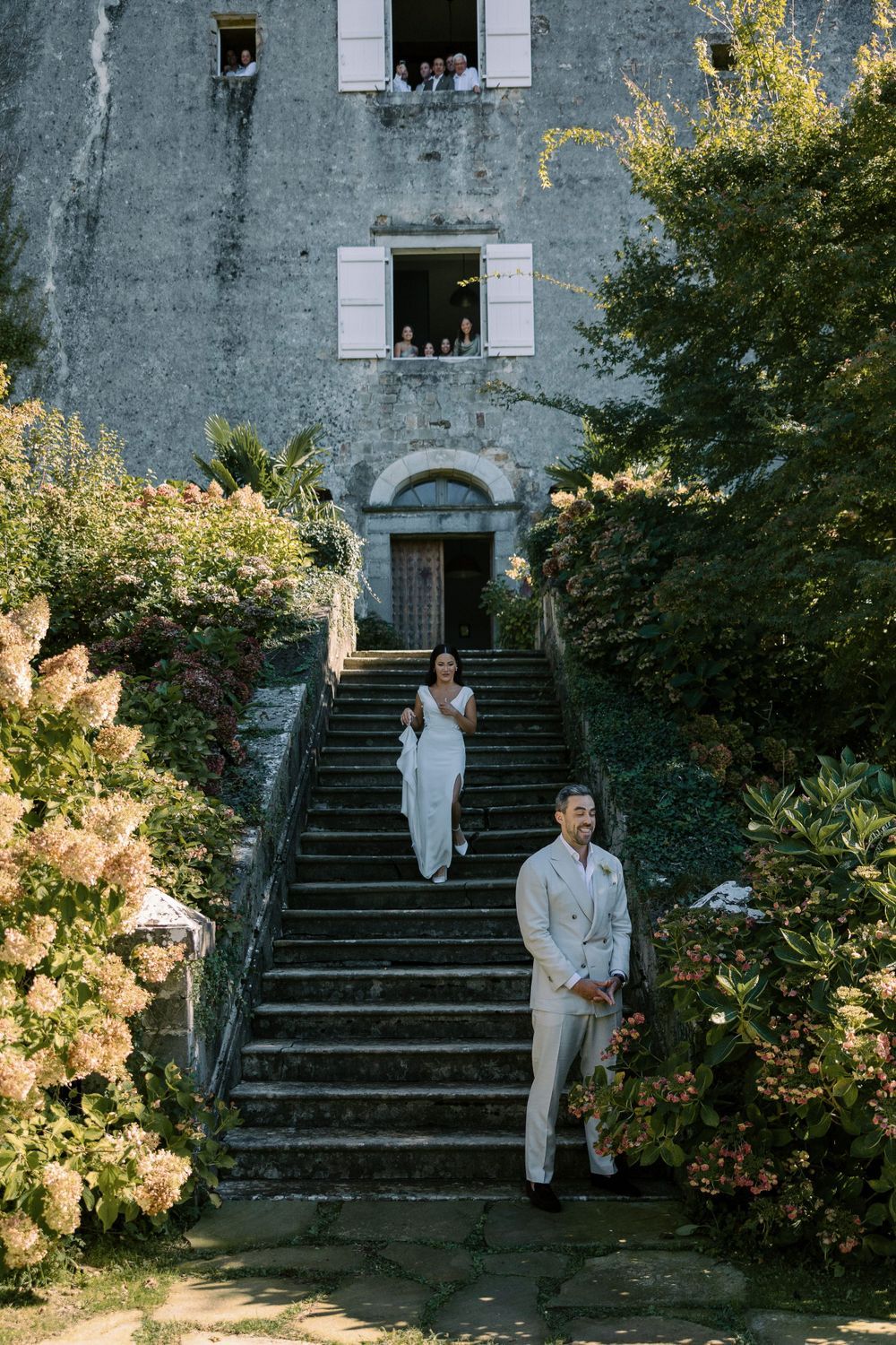 A wedding photoshoot on stone steps of a grey building surrounded by blooming hydrangeas.