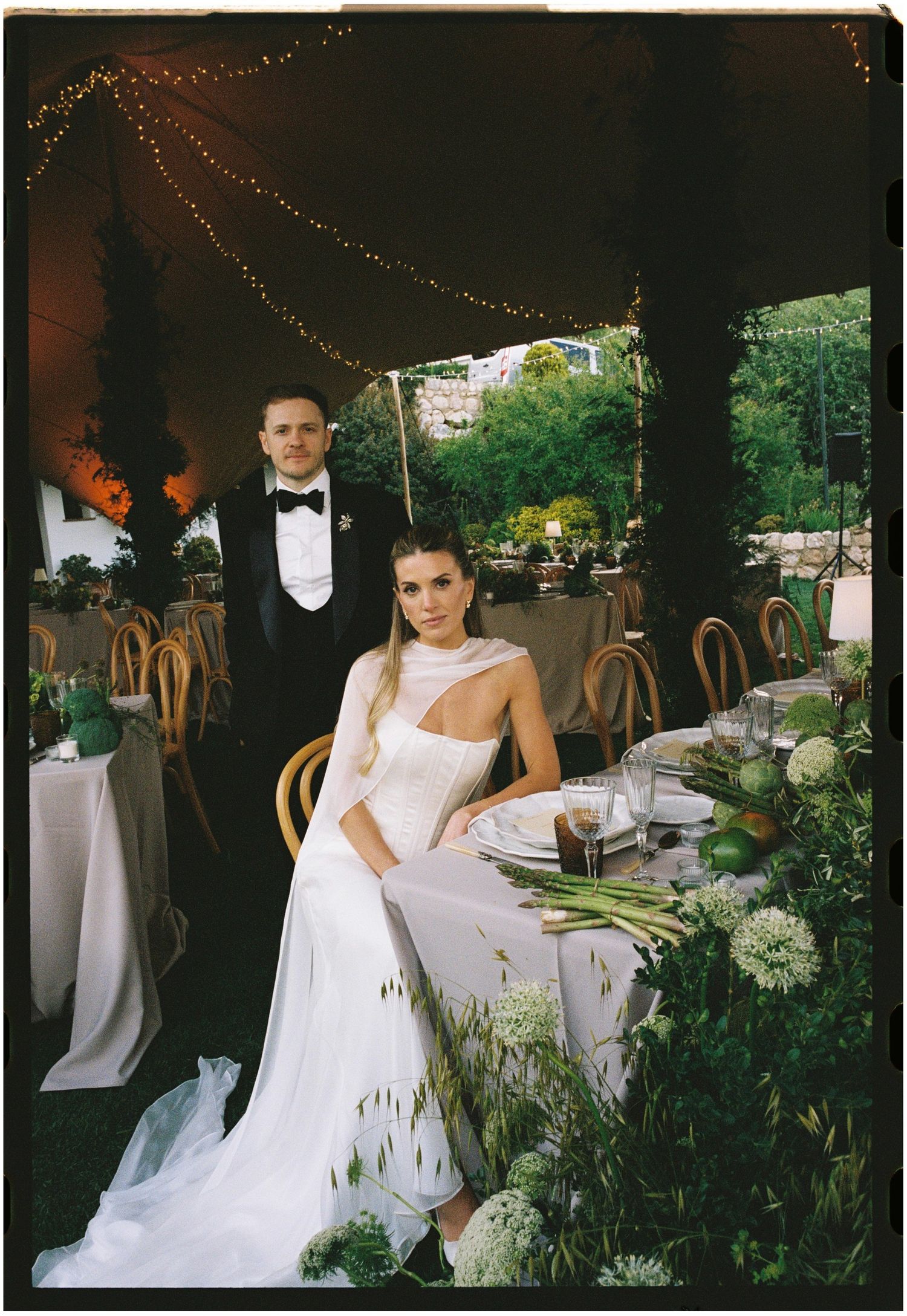 Elegant bride in white gown and groom in black tuxedo pose at floral-decorated wedding reception table under string lights.