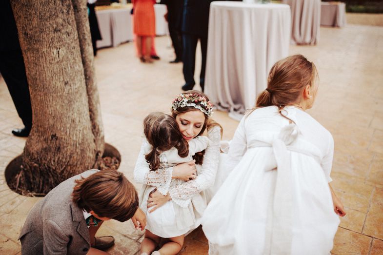 Young attendants in white dresses gathered around a tree trunk at an outdoor event.