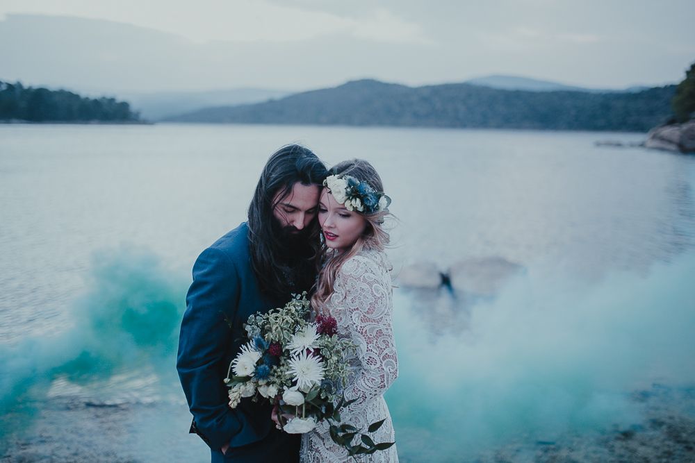 Romantic lakeside moment with turquoise smoke and white floral bouquet against mountain backdrop.