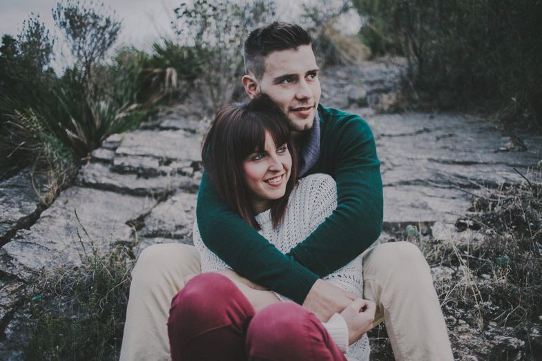 A couple embraces while sitting on rocky terrain with natural vegetation in the background.