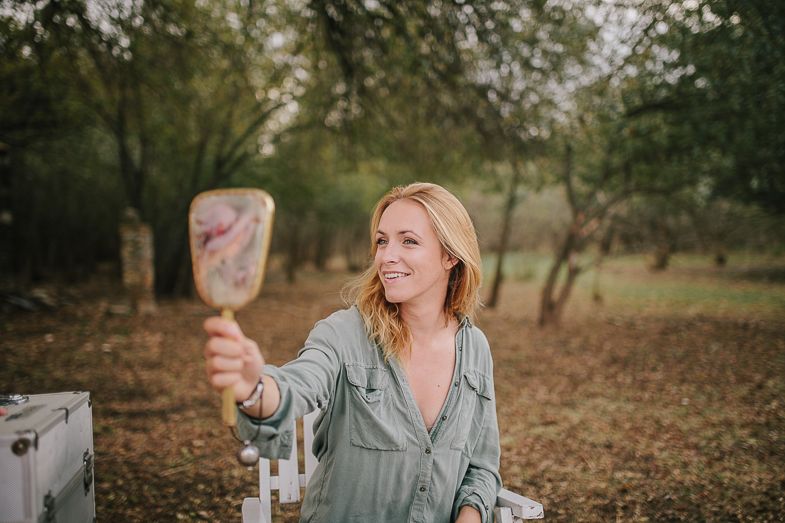 Person holding up a mirror during an outdoor makeup session with trees in background.