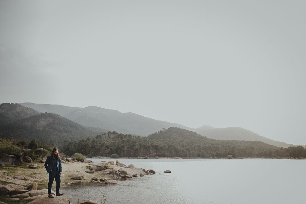 Lone figure walking along rocky shoreline with misty mountains in background.