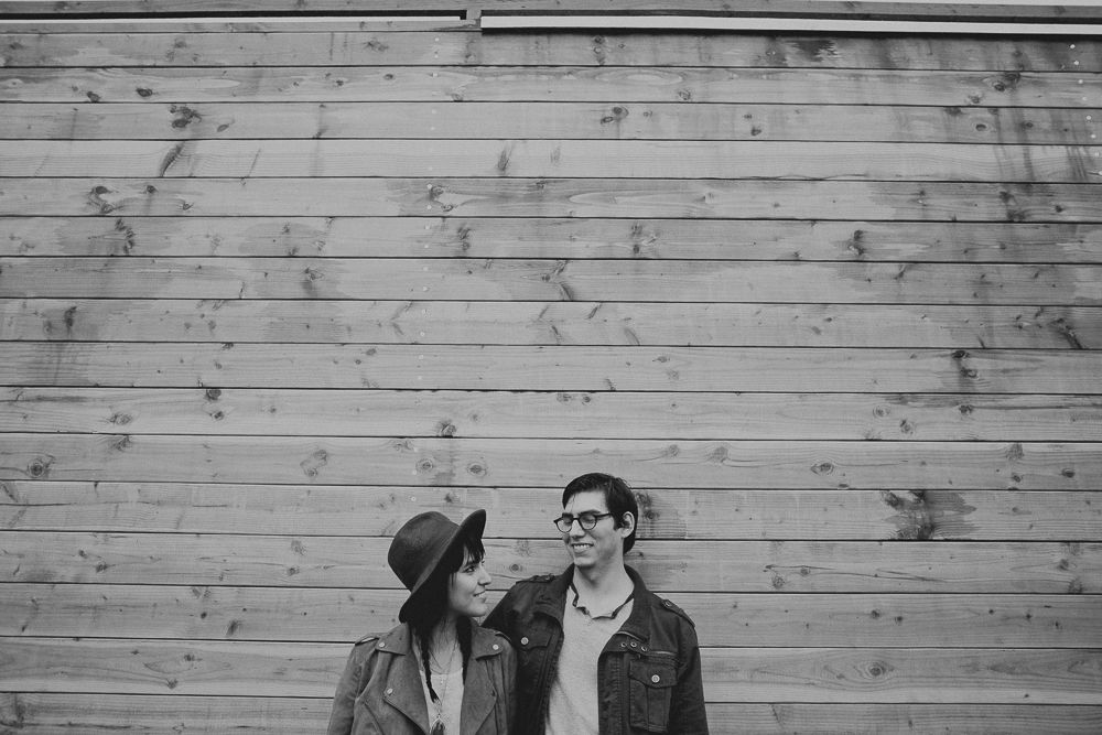 A romantic couple stands against a rustic wooden wall backdrop in black and white.