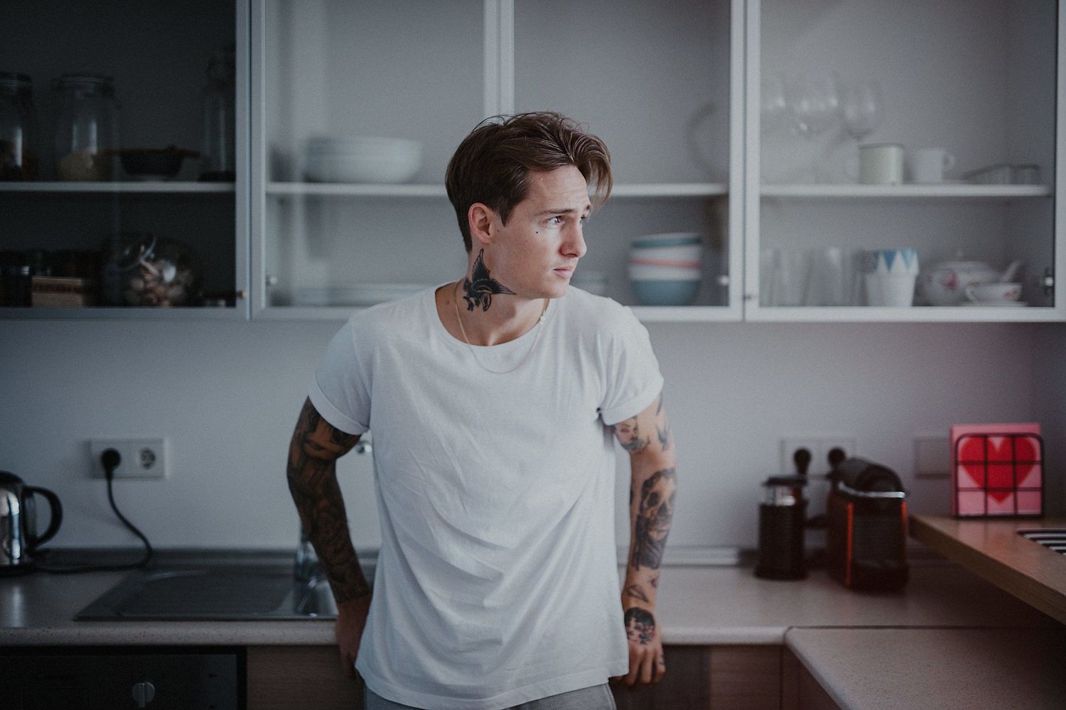 Man in white t-shirt with tattoos standing in modern kitchen.