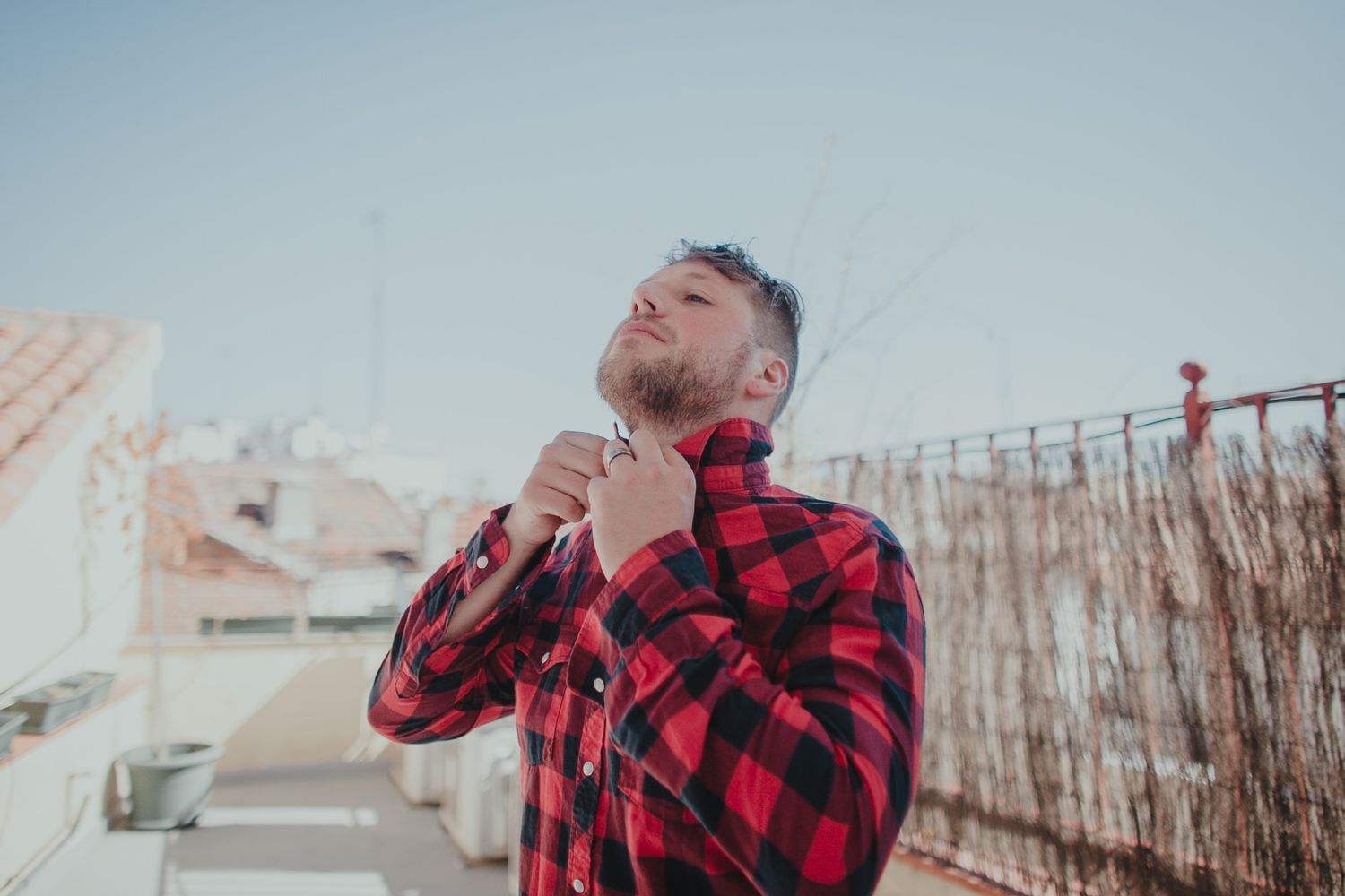 Person in red plaid shirt adjusting collar on sunny rooftop terrace.