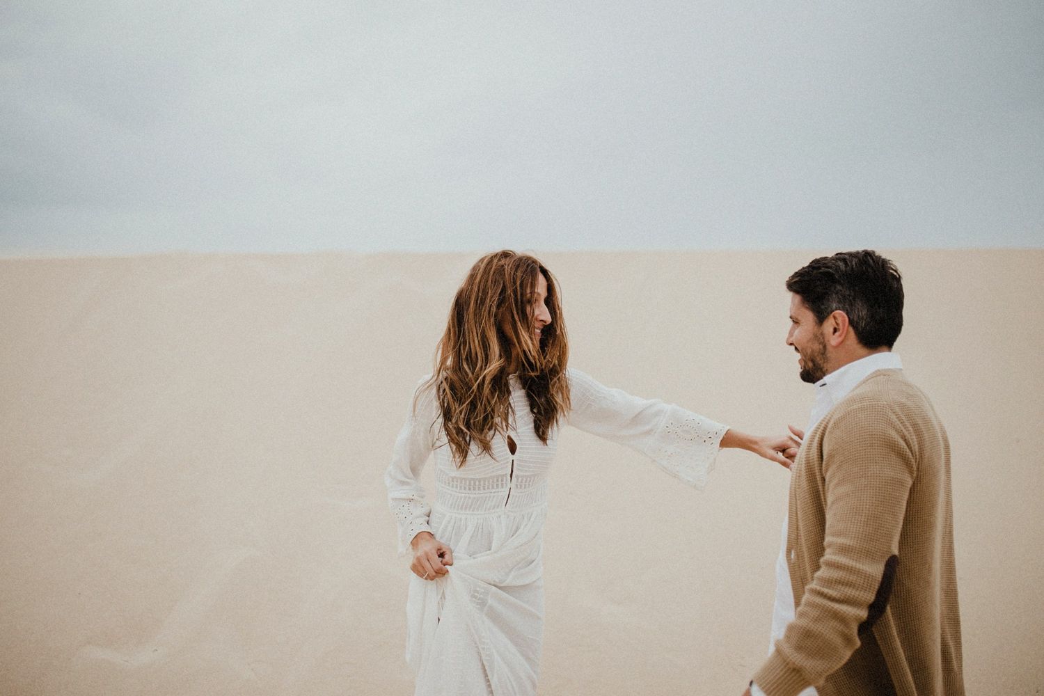 A couple in casual attire holds hands romantically while standing in a desert landscape against an overcast sky.