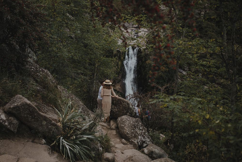 A figure stands near a cascading waterfall on a rocky forest trail.