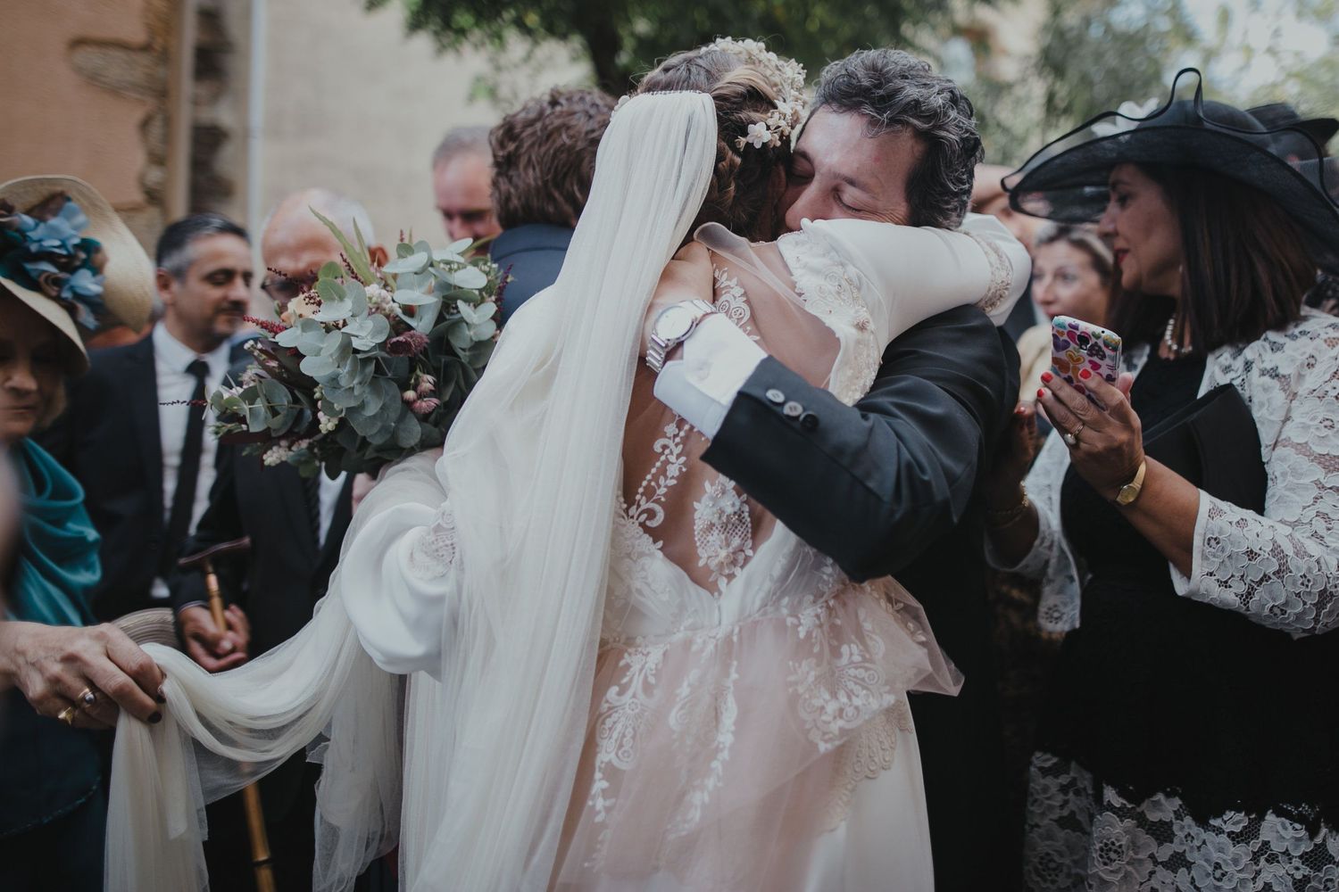 Wedding celebration with guests sharing emotional embraces outside a stone building.