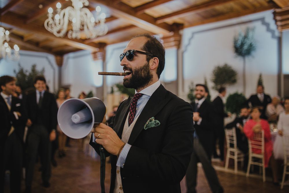 Wedding guest in sunglasses and suit jacket speaks into a megaphone at an indoor reception.