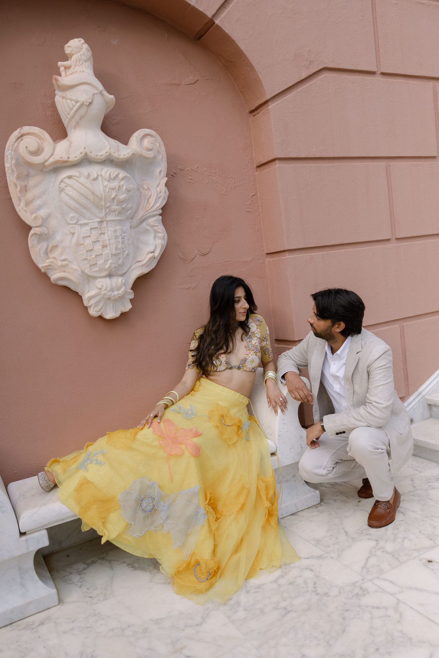 Elina seated in yellow beside Jag near a marble bench and decorative wall relief.