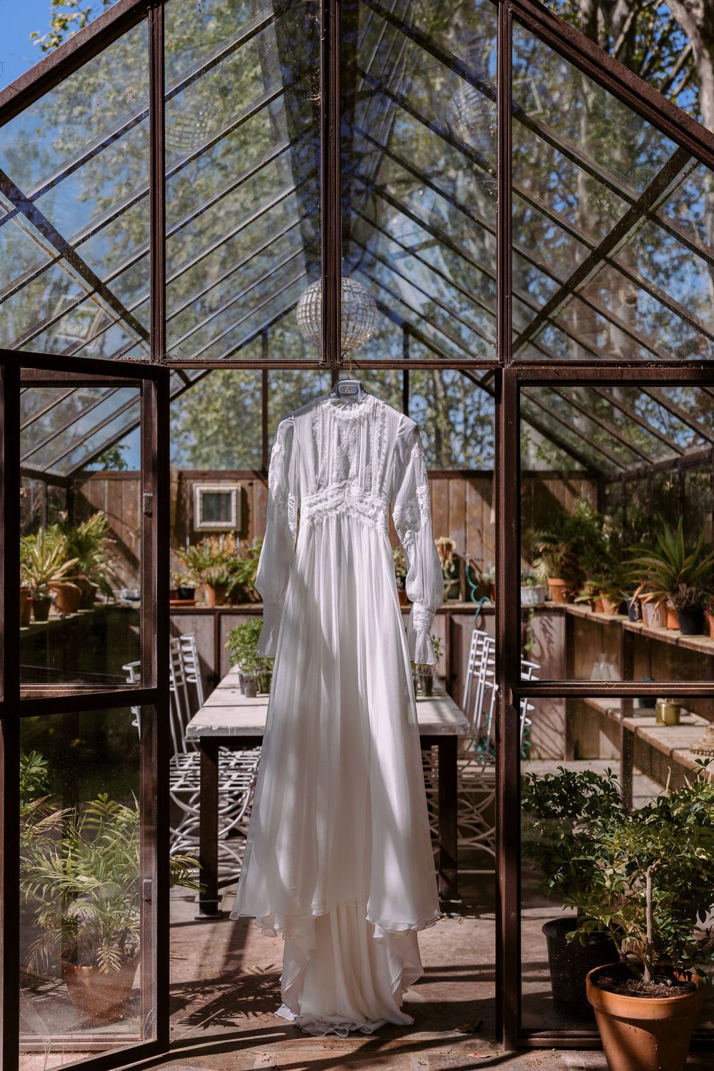 White wedding dress displayed in a sunlit greenhouse with glass walls and potted plants.