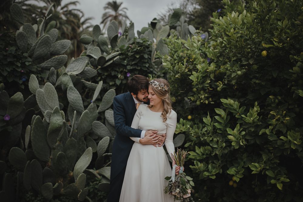 A wedding couple embraces in a tropical garden with palm trees and cacti.