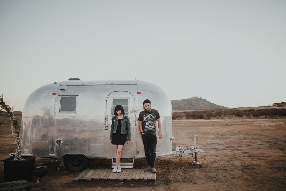Two people pose in front of a classic silver Airstream trailer in a desert setting.