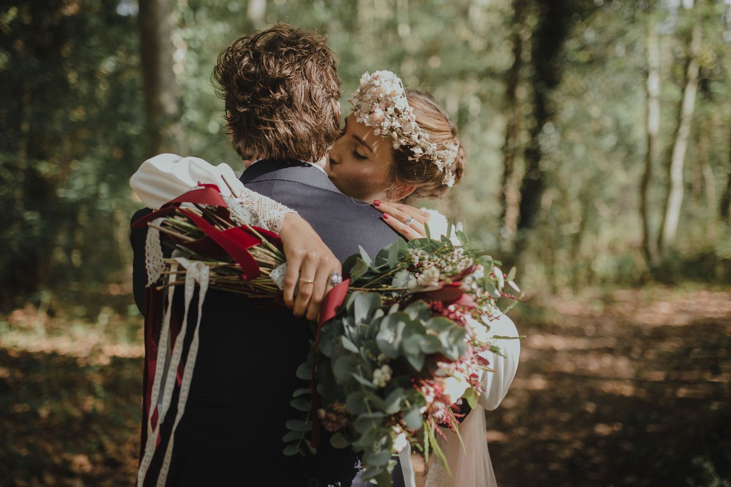 A tender embrace between newlyweds holding a bouquet with red ribbons in a forest setting.