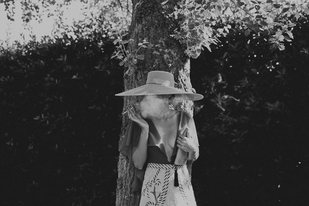 Black and white portrait of person in vintage dress and hat leaning against tree.