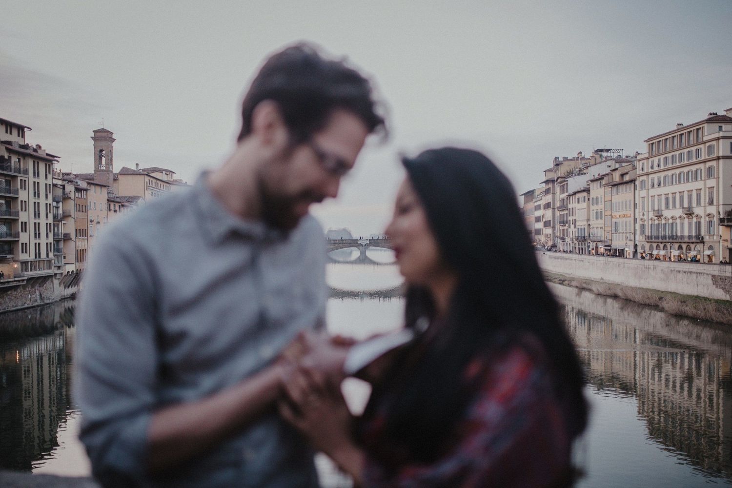 Couple shares a tender moment along the Arno River with historic buildings reflecting in the water.