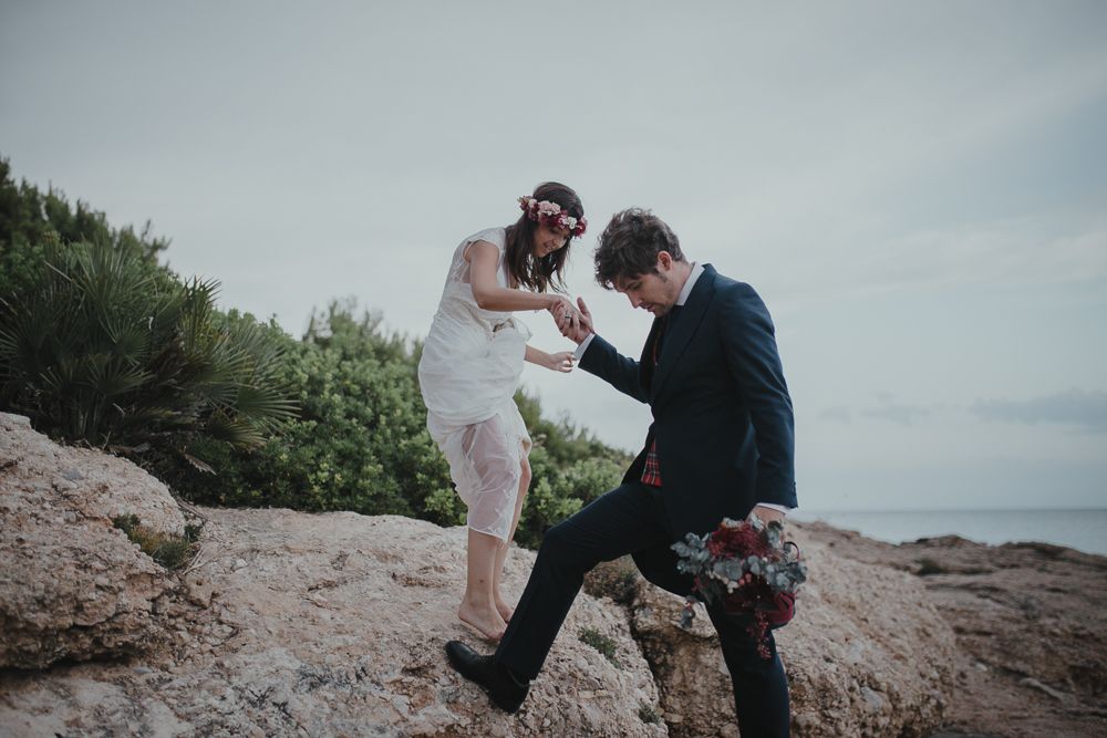 A couple having fun on coastal rocks in wedding attire.
