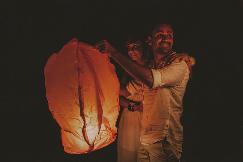 Three people light a paper lantern at night creating a warm orange glow.