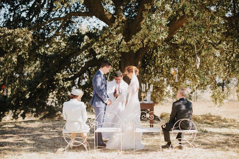Wedding ceremony taking place under a large tree with guests seated in white chairs.