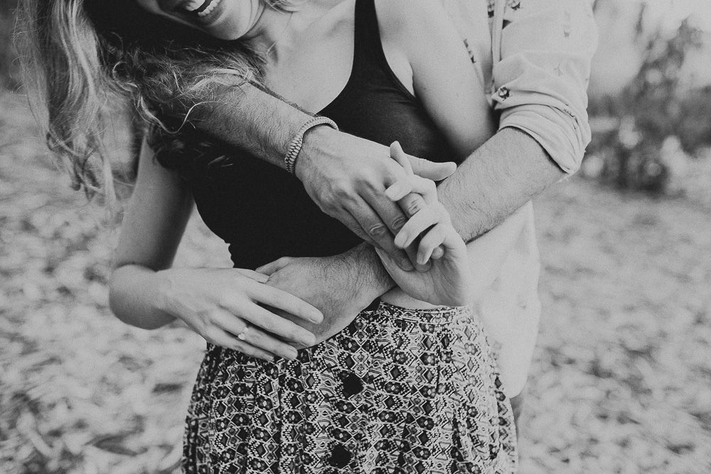 Black and white image of two people embracing with sunlight filtering through trees.