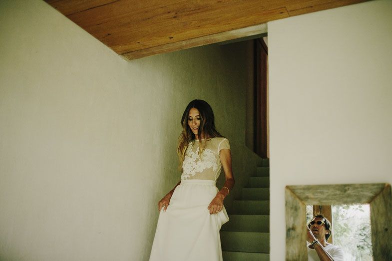 A figure in a delicate white wedding gown stands by a staircase in a rustic interior setting.