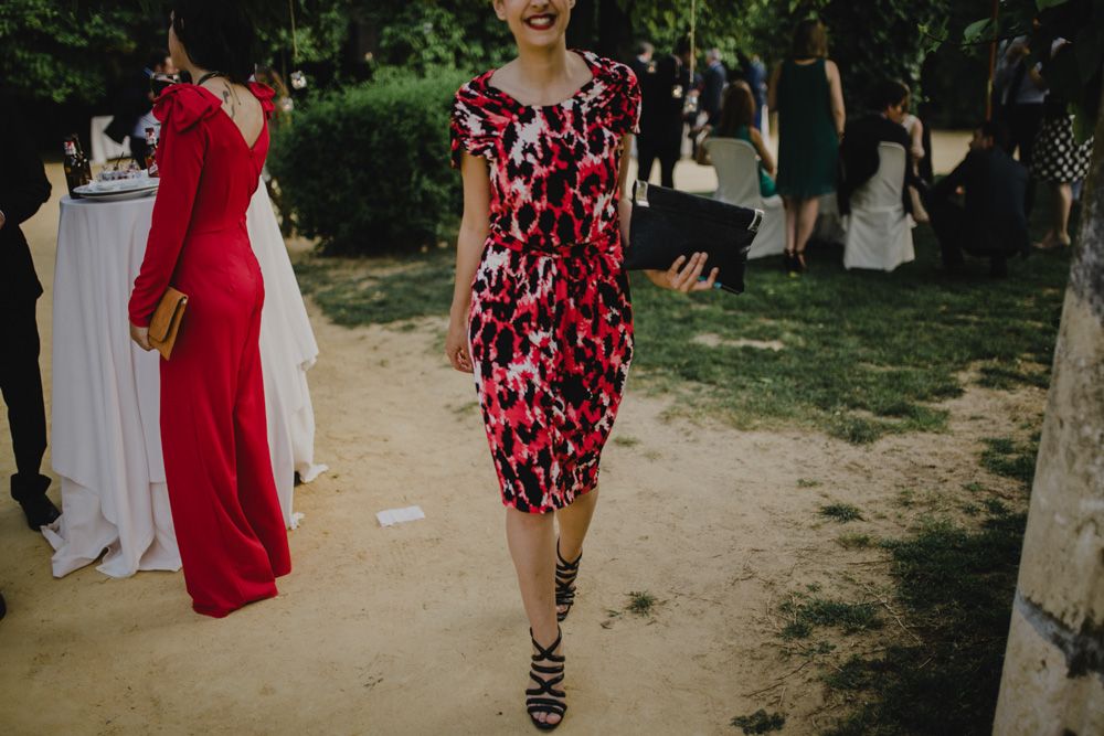 Guest in red floral dress walking on garden path during outdoor wedding reception.