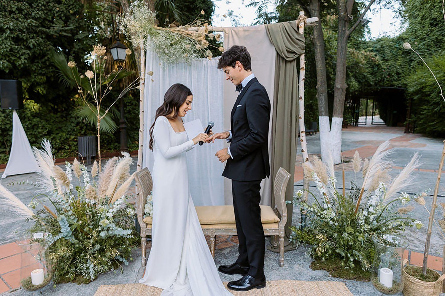 Couple exchanging rings during outdoor wedding ceremony under floral arch.