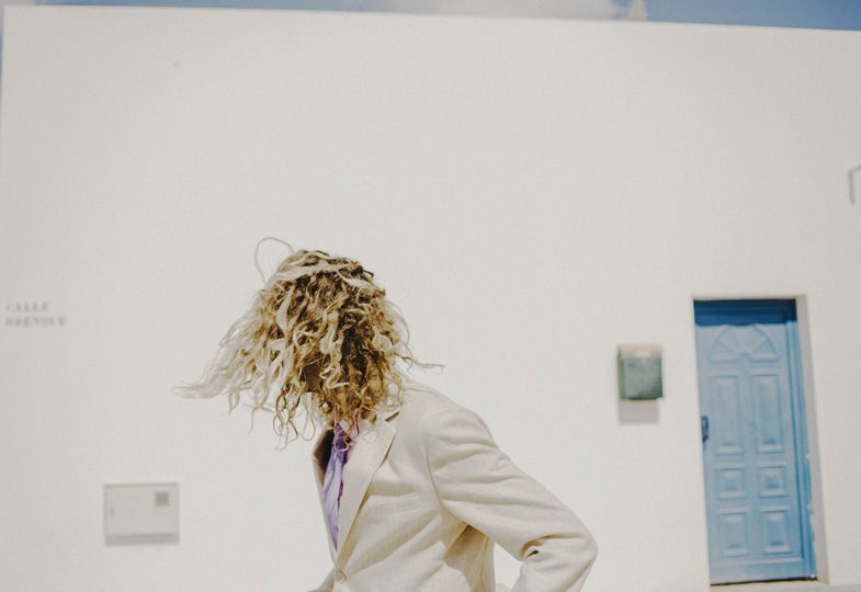 Person in white suit with curly hair against white wall with blue door.